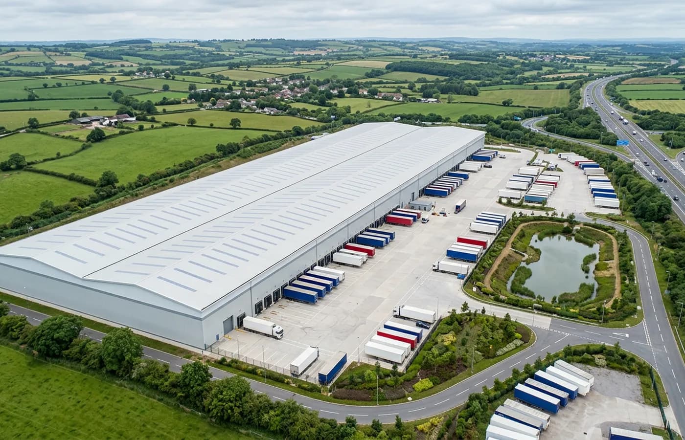 A large distribution warehouse beside a South West motorway junction, a long pale roof with rooflight strips and a loading dock apron