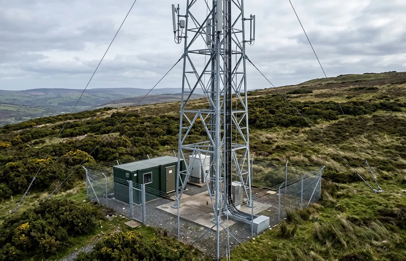 A galvanised steel lattice communications mast on a Devon hillside with antenna mounts and a cable tray, photographed from the air